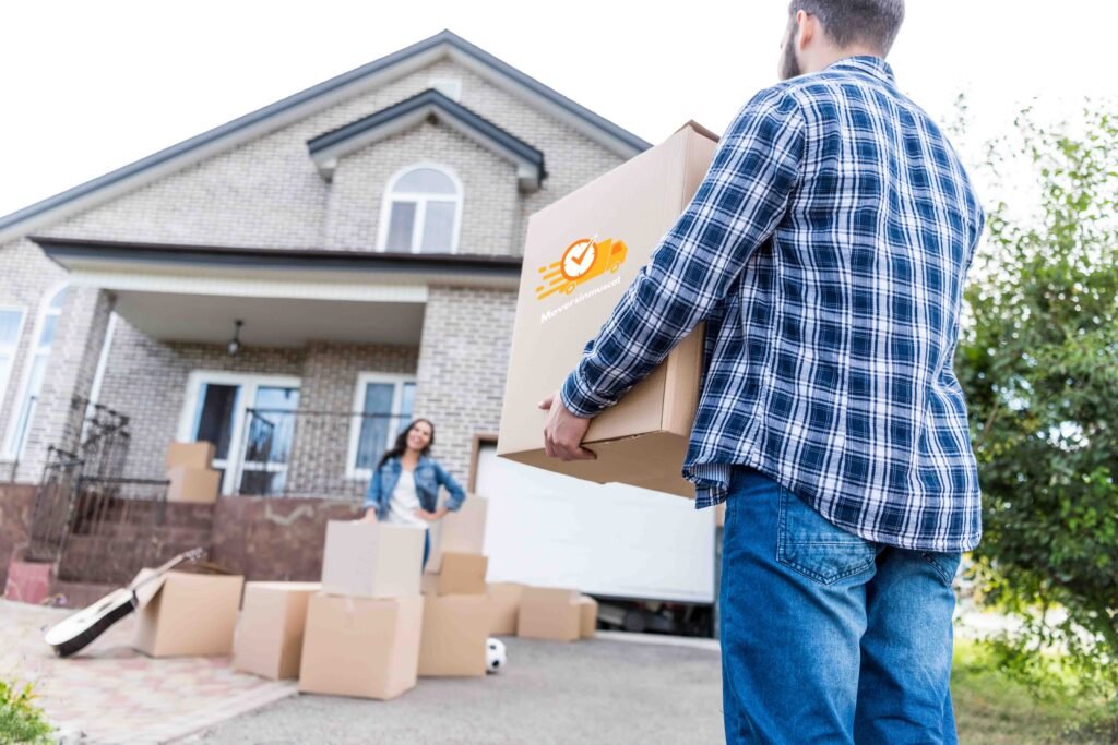 A man carrying a cardboard box toward a house, with a woman standing among several packed boxes, symbolizing house shifting services by Movers in Musca